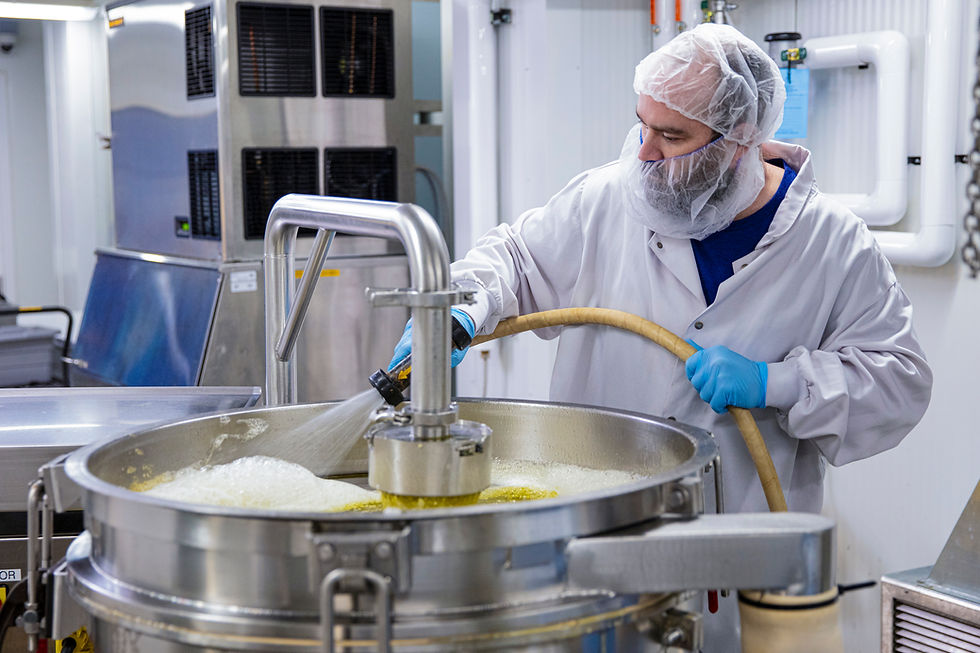 A photo of an Organigram team member at our Mocton site wearing a white labcoat, hair net and beard net, the worker is washing out a large metal drum with a hose