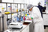 A photo of a team member wearing a lab coat and hair net, inspecing some colour swatches and diffeent package options, there are beekers with bright coloured liquids in the background