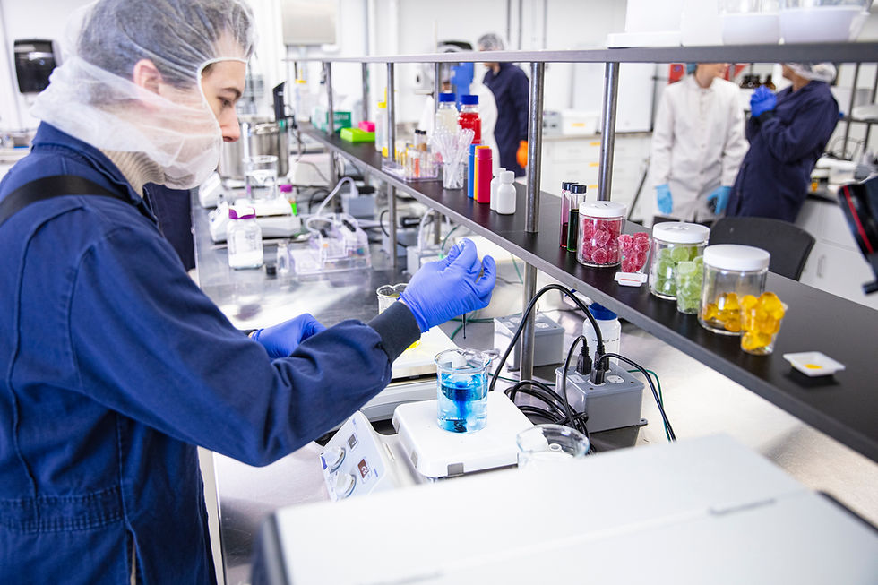 An image of a lab technician injecting some blue dye into a beeker, there are brightly coloured gummies in jars in the background as well as other team members working